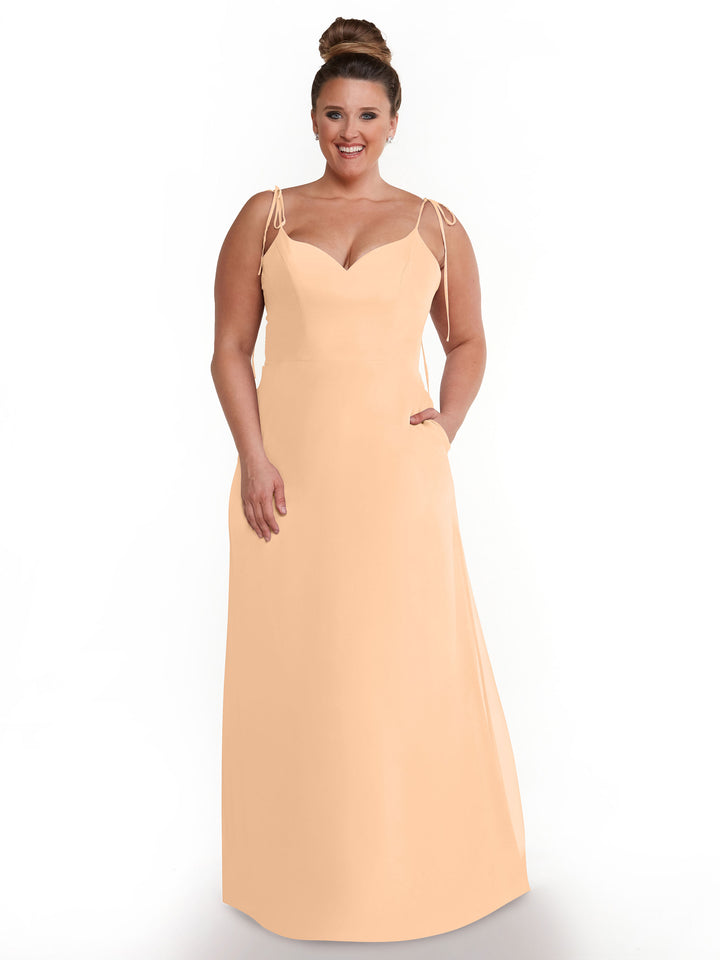 A woman models Avery Austin dress 80144, a peach flowy chiffon style with spaghetti straps and a V-neck. She stands against a white background, smiling with her hair in a high bun and one hand in the dress pocket.