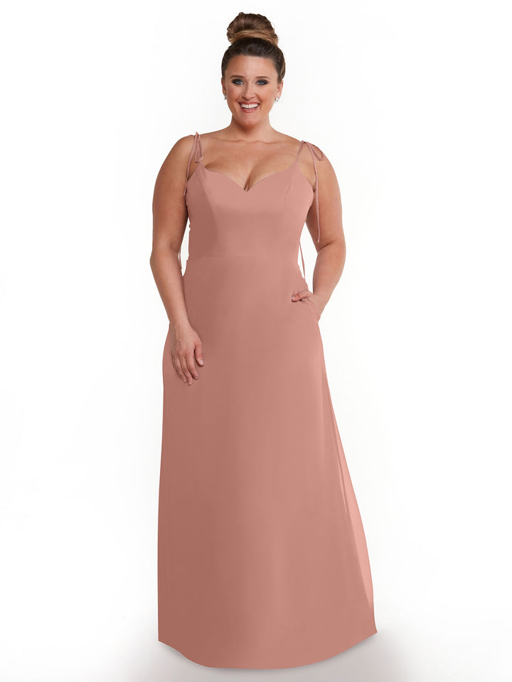 A woman with her hair in a bun wears the Avery Austin 80144 sleeveless rose chiffon V-neck dress with thin straps and pockets, standing against a plain white background.