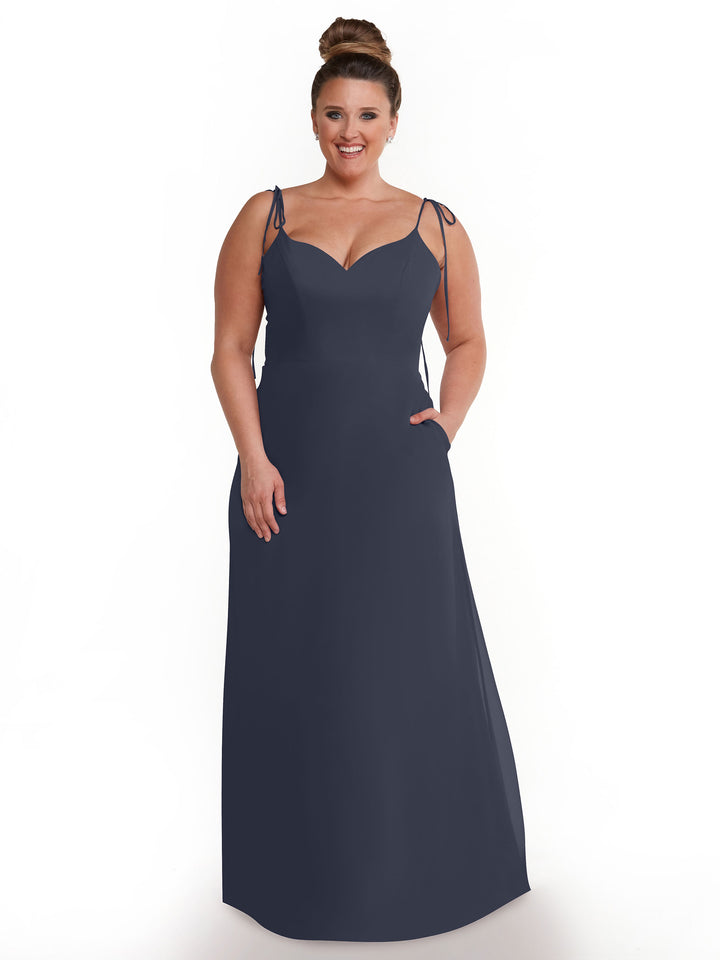 A woman with brown hair in a bun is modeling Avery Austin’s 80144, a sleeveless navy blue flowy dress featuring a V-neck, thin straps, and pockets, against a plain white background.