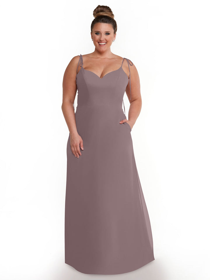 A woman with her hair in a bun stands against a white background, wearing Avery Austin 80144—a sleeveless mauve chiffon dress with thin straps, a sweetheart neckline, and flowy pockets.