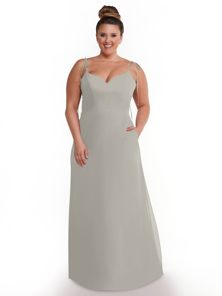 A woman with brown hair in a top bun models Avery Austin’s 80144: a light gray, sleeveless, flowy chiffon dress with thin straps and pockets, standing and smiling against a white background.