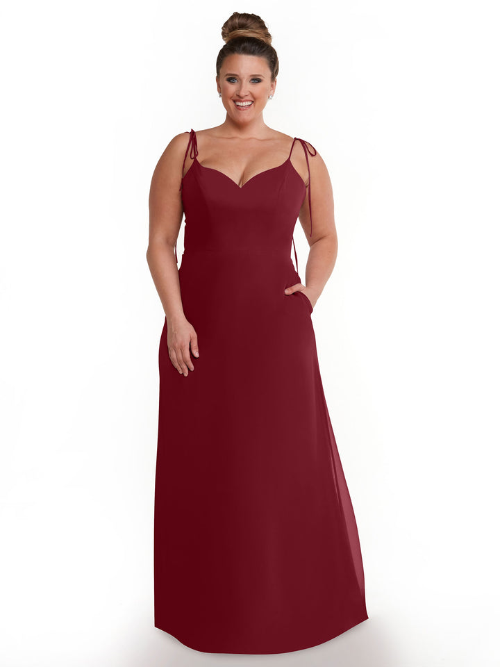 A woman models the Avery Austin TH-80144, a sleeveless burgundy chiffon V-neck dress with shoulder ties, standing against a white background and smiling with her hand in her pocket and hair in a neat bun.