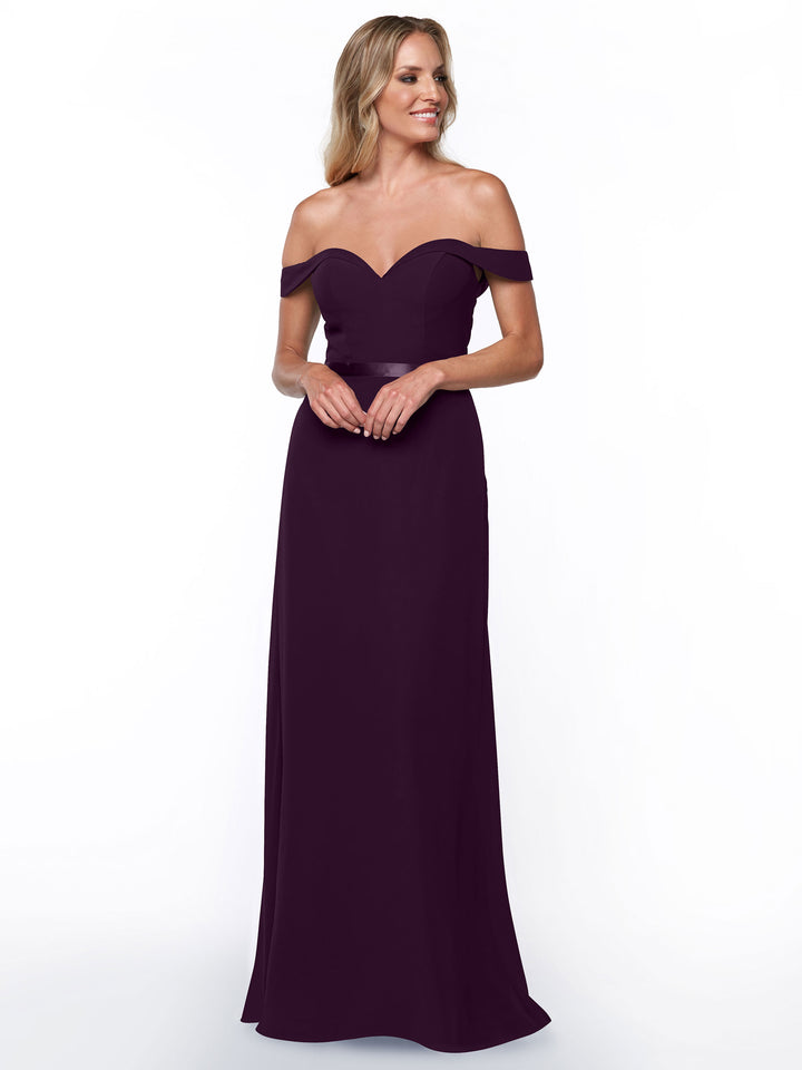 A woman in Avery Austin’s 80145, an elegant off-the-shoulder, floor-length deep purple chiffon bridesmaid dress with a sweetheart neckline, stands against a white background, smiling softly with her hands clasped.