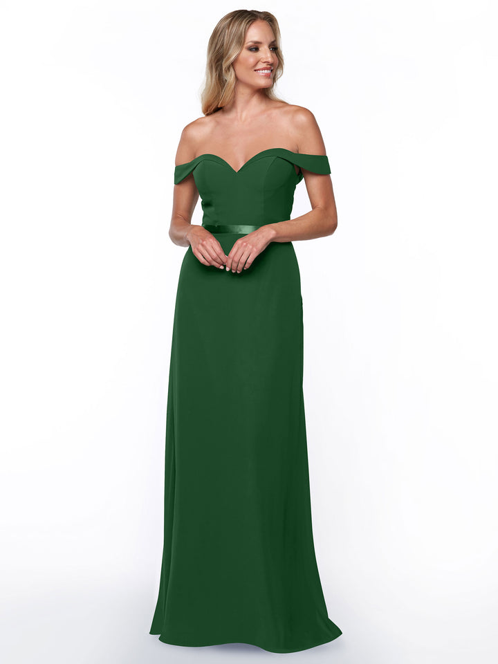 A woman stands smiling against a plain white background, wearing Avery Austin’s 80145: an elegant, floor-length emerald green bridesmaid dress with a fitted bodice and subtle sweetheart, off-the-shoulder neckline.