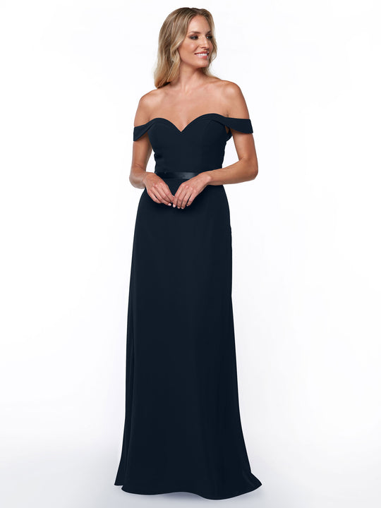 A woman stands and smiles against a white background, wearing Avery Austin’s TH-80145 bridesmaid dress in Midnight—a long, off-the-shoulder gown with a sweetheart neckline.