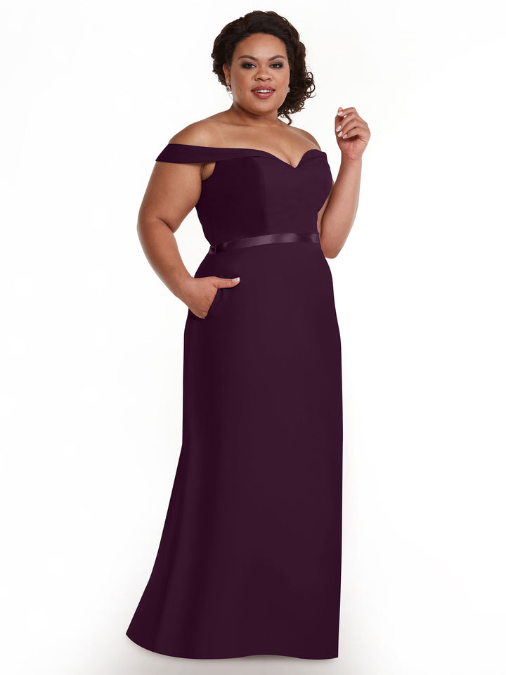 A woman poses confidently in the Avery Austin 80145, an off-the-shoulder, floor-length deep purple bridesmaid dress with a satin waistband and pockets, set against a white background.