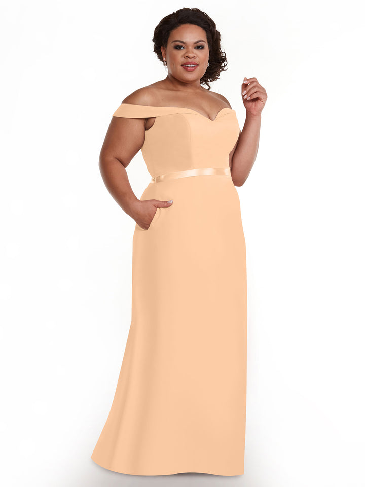 A woman in Avery Austin’s 80145, an off-the-shoulder, floor-length peach gown with a satin waistband and side pockets, smiles against a white background.