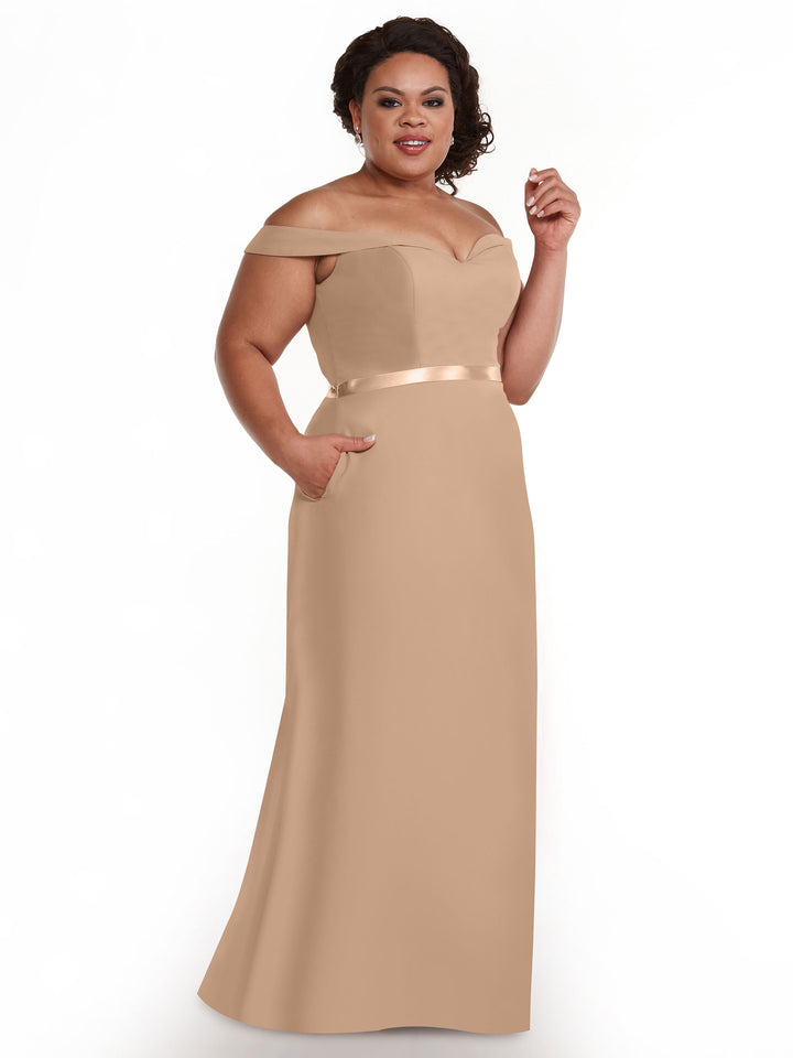 A woman wearing the Avery Austin 80145, a floor-length tan off-the-shoulder bridesmaid dress with a satin waistband and pockets, smiles against a plain white background.