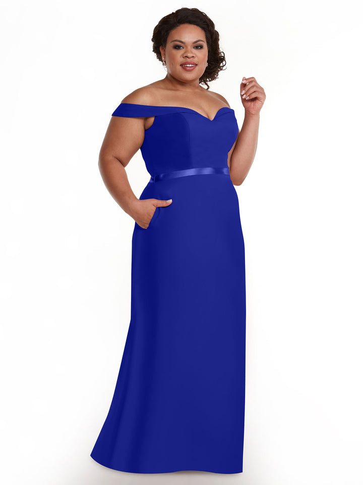 A woman smiles with one hand in her pocket, modeling Avery Austin’s 80145: a royal blue, off-the-shoulder bridesmaid dress featuring a sweetheart neckline, satin waistband, and floor-length A-line silhouette against a white background.