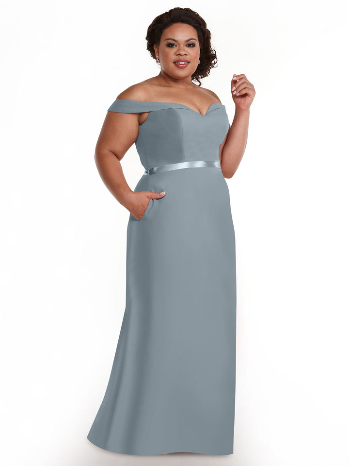 A woman wears Avery Austin style 80145, a light blue off-the-shoulder bridesmaid dress with a satin waistband, smiling as she poses with one hand in her pocket against a plain white background.