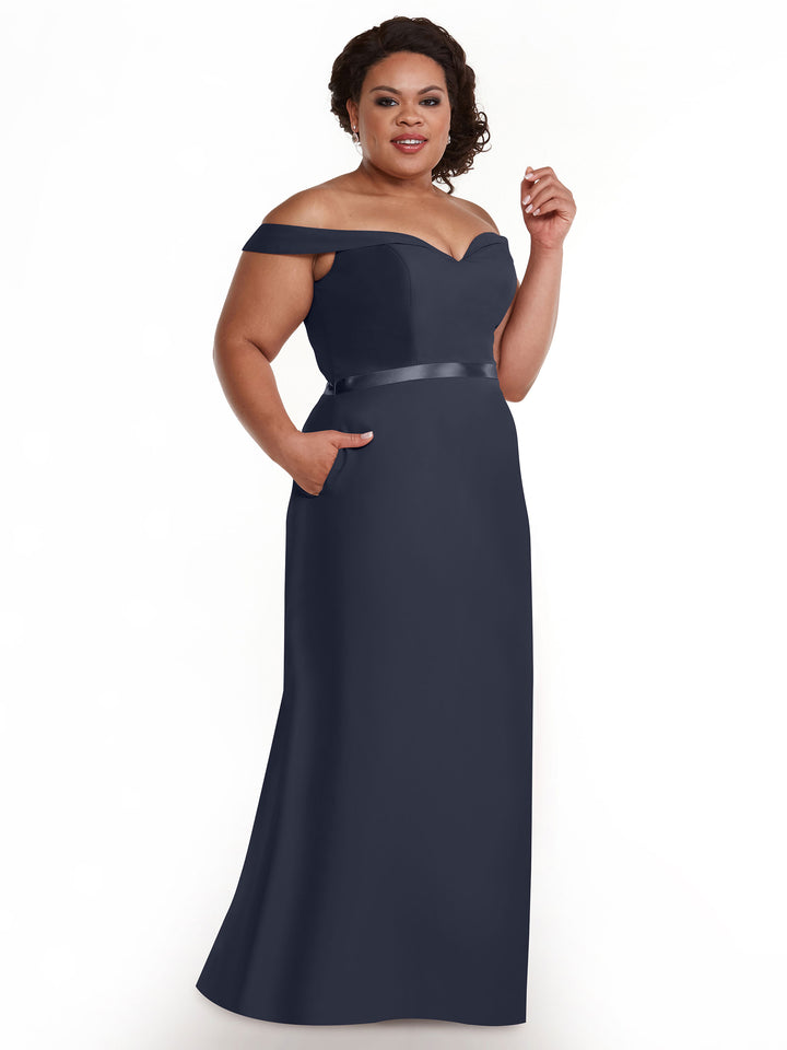 A woman models the Avery Austin 80145, a floor-length navy blue off-the-shoulder bridesmaid dress with a satin waistband and pockets, smiling against a plain white background.