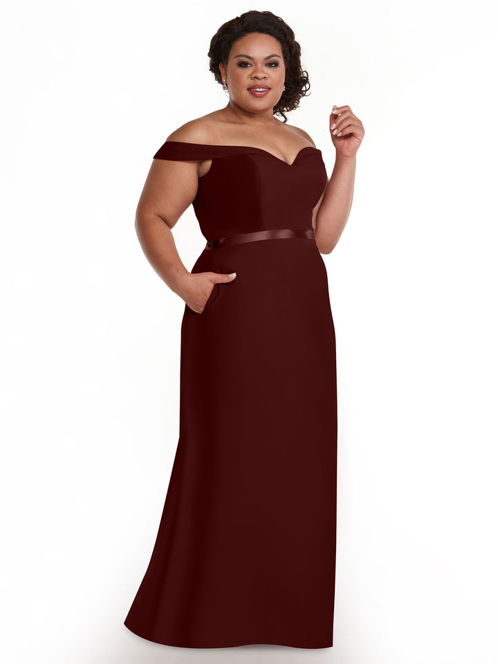 A woman smiles against a white background, wearing Avery Austin’s 80145—a dark burgundy, off-the-shoulder A-line bridesmaid gown with a satin waistband, pockets, and floor-length skirt.
