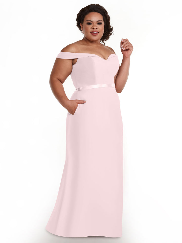 A woman in the Avery Austin TH-80145 bridesmaid dress in Rose Petal, featuring an off-the-shoulder neckline, satin waistband, and side pockets, smiles against a white background.
