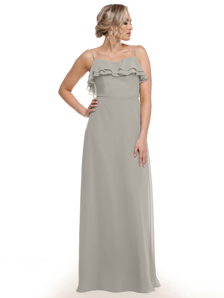 A woman stands against a white background, hand on her hip and hair in an updo, wearing Avery Austin style 80146—a long light gray chiffon A-line dress with tiered ruffles on the bodice.