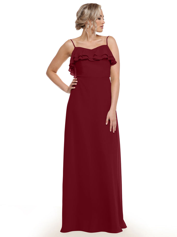 A woman with light hair styled up stands with one hand on her hip, wearing Avery Austin’s 80146—a long, sleeveless chiffon A-line dress featuring thin straps and tiered ruffles across the chest. She wears no visible accessories.