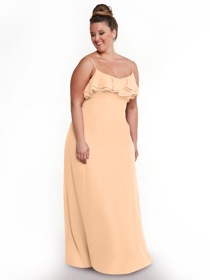 A woman models the Avery Austin 80146: a long, sleeveless, peach chiffon A-line dress with thin straps and tiered ruffles on the chest, standing against a plain white background, smiling and looking slightly down.