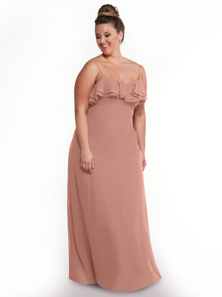 A woman wears the Avery Austin 80146 blush pink chiffon A-Line dress with tiered ruffles at the neckline, standing on a plain white background and smiling as she looks down to her left.