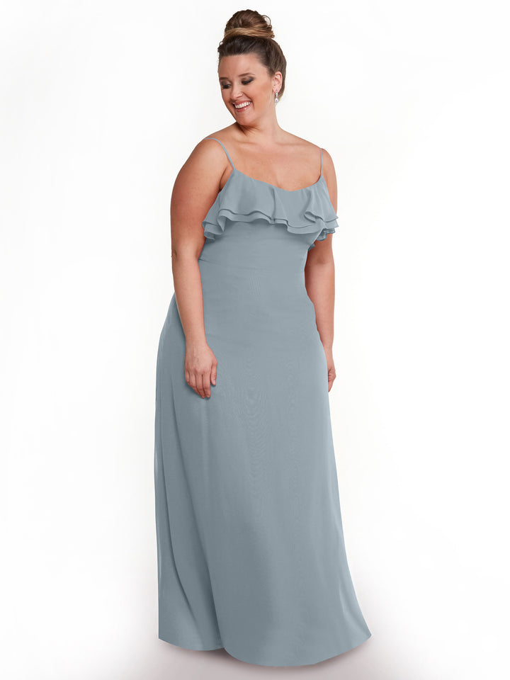 A woman in the Avery Austin 80146 light blue chiffon A-line dress with thin straps and a ruffled neckline stands against a white background, smiling and looking down.