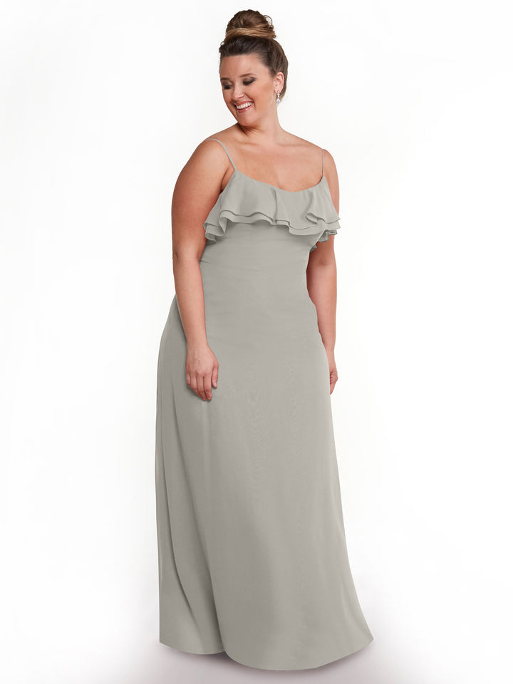 A woman wearing Avery Austin’s 80146 light gray chiffon A-Line dress with thin straps and a ruffled neckline stands smiling against a plain white background.