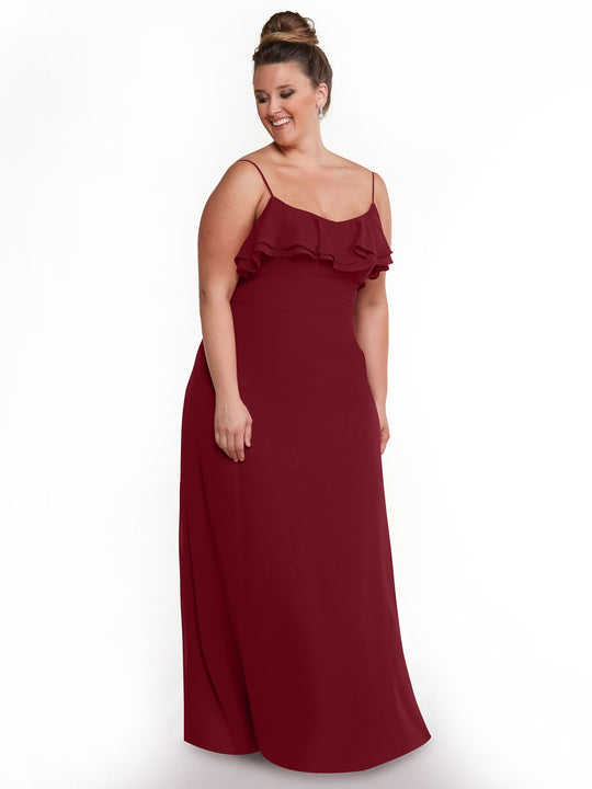 A woman in the Avery Austin TH-80146 sleeveless floor-length deep red chiffon A-line dress with tiered ruffles at the bust stands against a white background, smiling while looking down to her right.