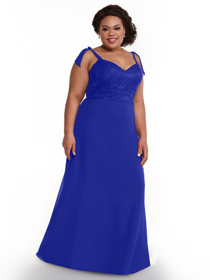 A woman stands smiling against a plain white background wearing Avery Austin’s royal blue chiffon dress 80149, featuring a floral lace bodice and self-tie straps.