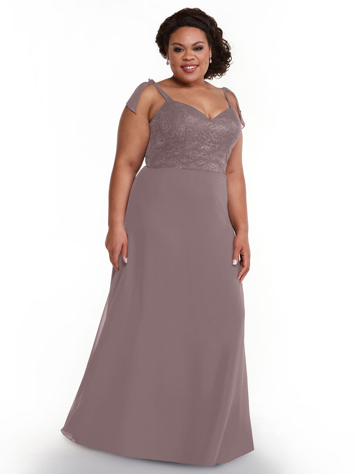 A woman in Avery Austin’s 80149 mauve, floor-length chiffon dress with floral lace bodice and self-tie straps stands smiling against a plain white background.