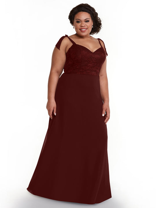 A woman in an Avery Austin TH-80149 floor-length merlot chiffon dress with a floral lace bodice and self-tie straps stands against a plain white background, smiling slightly.