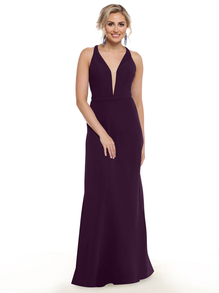 A woman with blonde hair in an updo wears Avery Austin’s 80150—a sleeveless, floor-length deep purple gown with a plunging neckline. She stands against a white background, smiling slightly, hands relaxed in front of her.