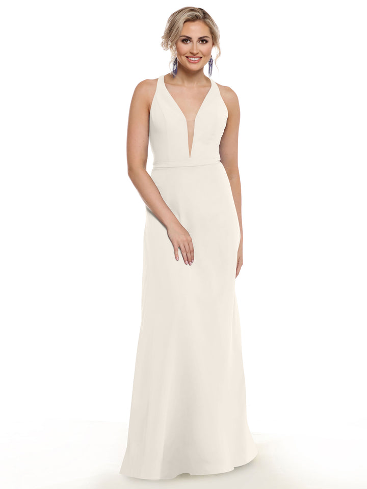 A woman with styled blonde hair and drop earrings stands against a plain white background, smiling with one hand on her hip, wearing Avery Austin’s 80150—a sleeveless, floor-length white gown with a plunging neckline.