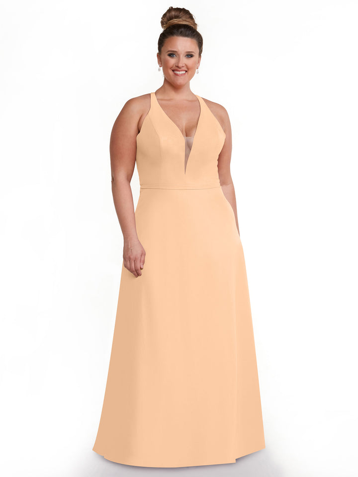 A woman with her hair in a bun smiles in front of a white background, wearing the Avery Austin 80150—a sleeveless, peach floor-length gown with a plunging neckline.