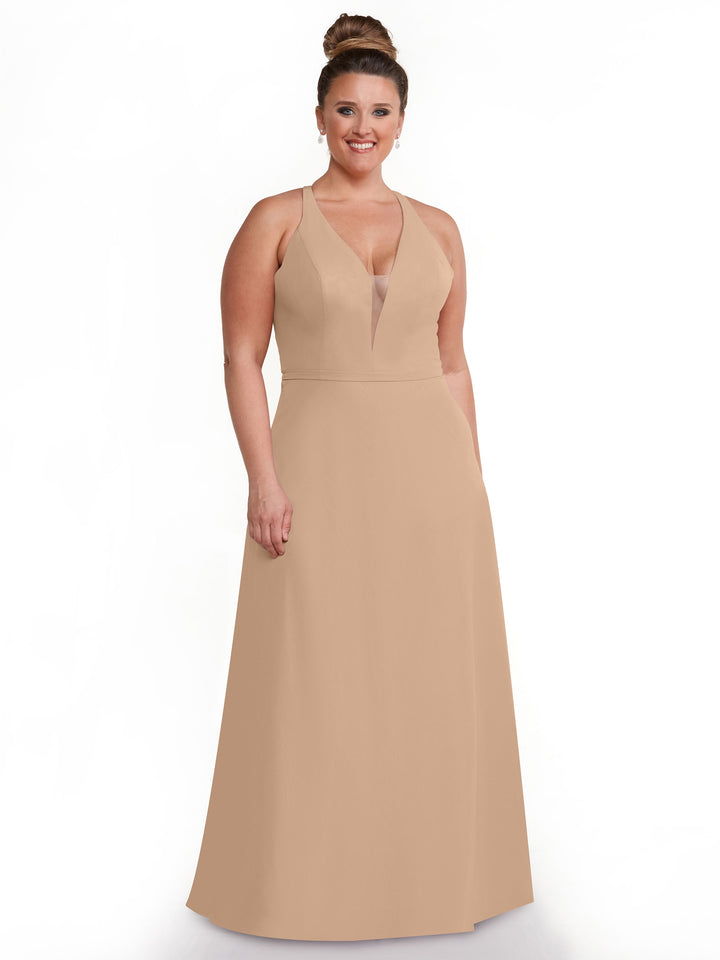 A woman with her hair in a bun stands and smiles against a plain white background, wearing Avery Austin’s 80150: a sleeveless, floor-length light beige dress with geometric cut-outs and a mesh insert.