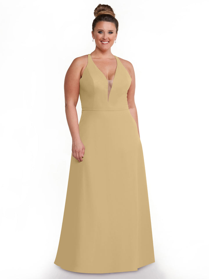 A smiling woman with light skin and brown hair in a bun models Avery Austin’s 80150: a sleeveless, V-neck, floor-length beige gown with pockets, posed against a plain white background.