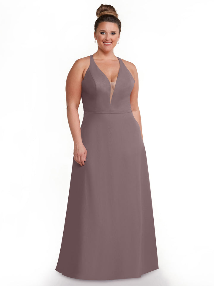 A woman with her hair in a high bun smiles in front of a white background, wearing Avery Austin’s mauve A-line gown (style 80150) featuring a plunging neckline and side pockets.