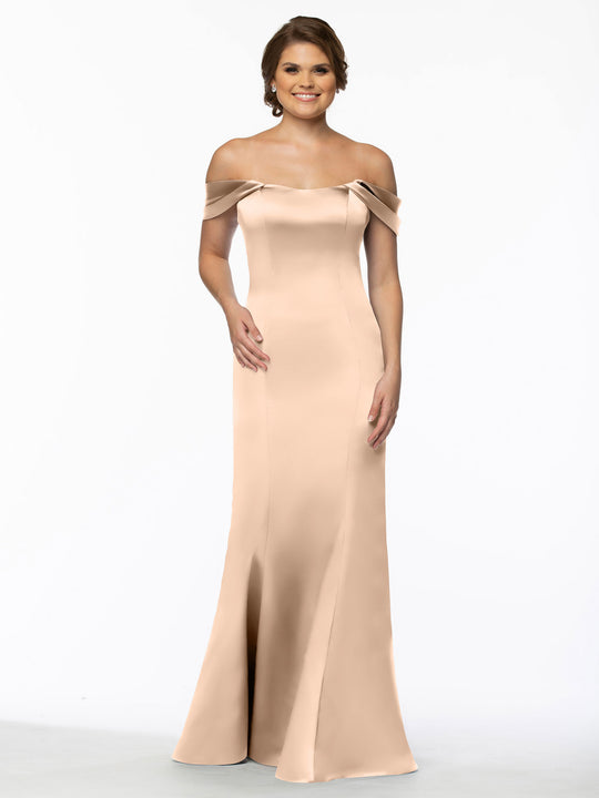A woman in a floor-length, off-the-shoulder Avery Austin 80151 dress in blush stands against a plain white background, smiling with one hand by her side and the other touching her waist.