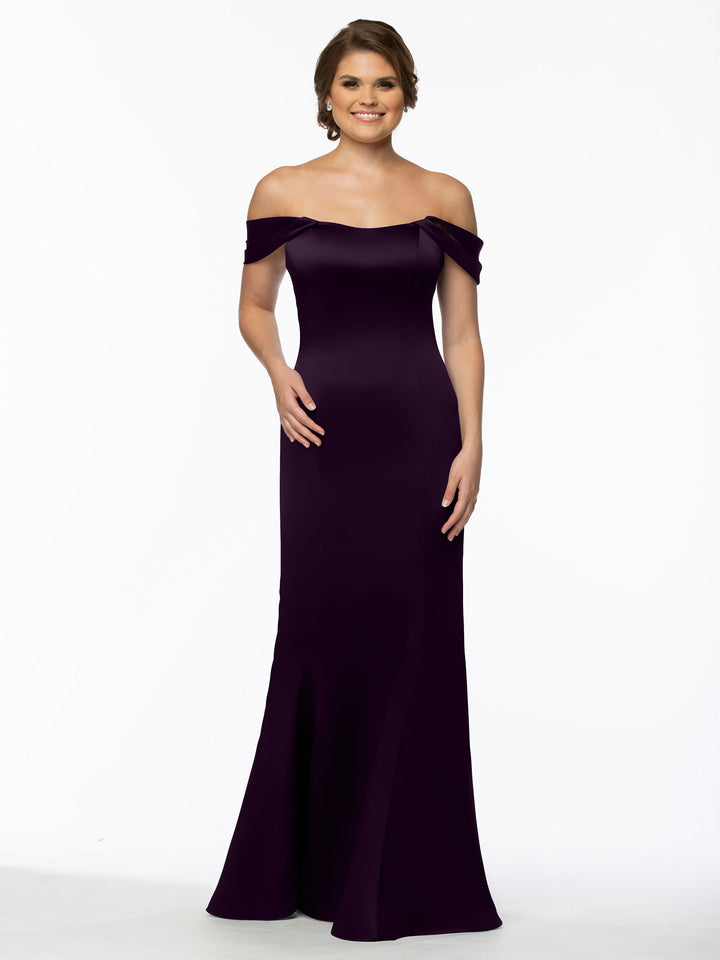A woman poses against a plain white background, smiling in the Avery Austin 80151 floor-length, off-the-shoulder fitted dress in dark purple with one hand on her hip and the other relaxed by her side.