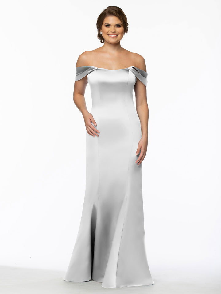 A woman smiles against a plain white background, wearing Avery Austin’s 80151—a silver satin off-the-shoulder gown with a fitted bodice, convertible straps, and a gently flared hem for an elegant finish.