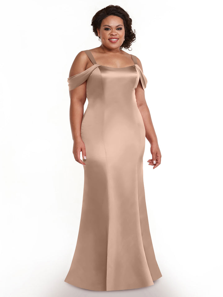 A woman stands smiling against a plain white background, wearing Avery Austin’s 80151—a floor-length, light brown fitted dress with off-the-shoulder straps.