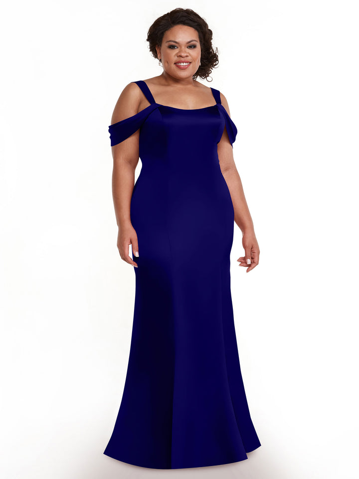 A woman wearing Avery Austin’s 80151 fitted, floor-length, off-the-shoulder dress stands against a white background, smiling with loose curls in her hair.