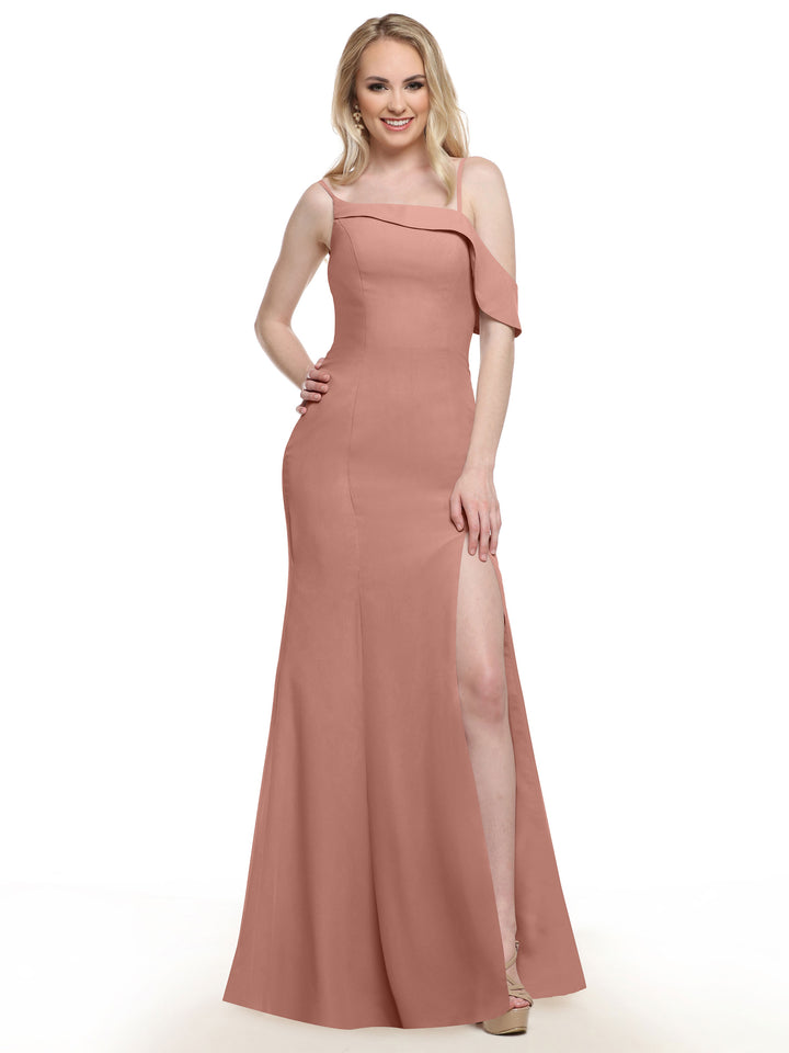 A woman stands smiling against a plain white background, wearing Avery Austin’s 80153 gown in Dusty Rose—a floor-length dress featuring a high slit and an asymmetrical ruffle sleeve.