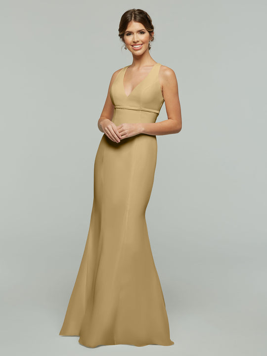 A woman in the Avery Austin 80155, a sleeveless, floor-length fit and flare dress in champagne, stands against a light gray background, smiling with her hands clasped in front.