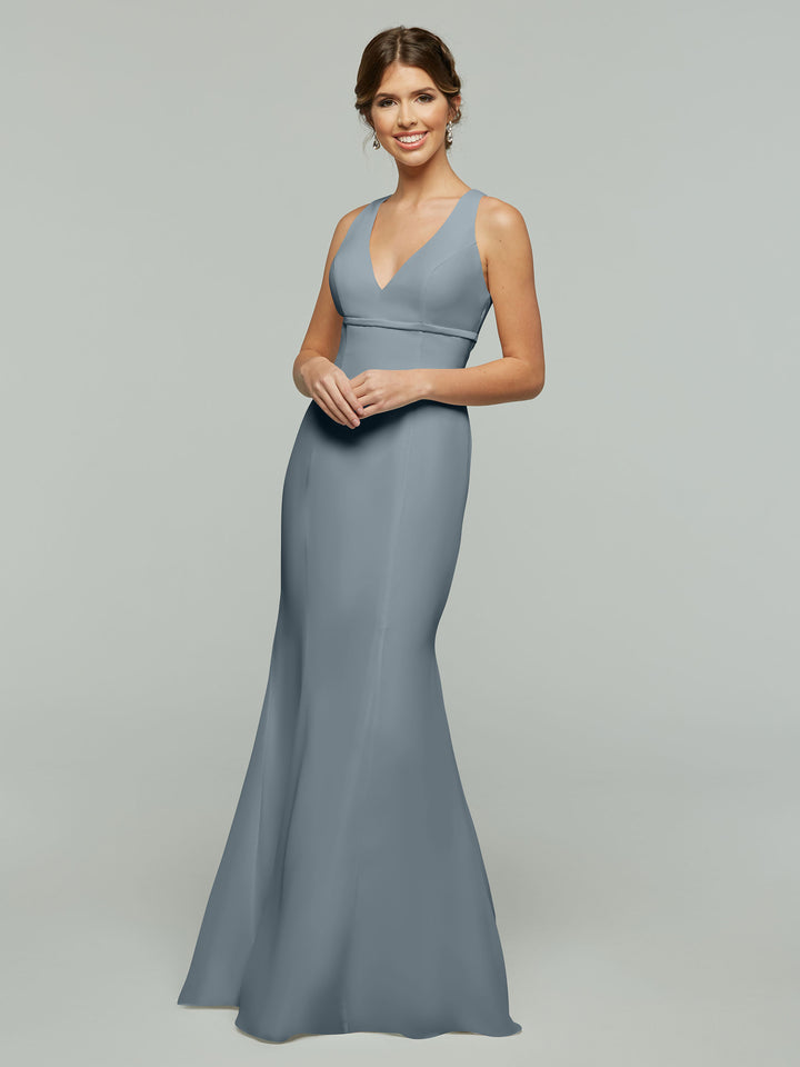 A woman smiles against a light gray backdrop, wearing Avery Austin’s 80155: a sleeveless, floor-length chiffon dress in dusty blue, featuring a V-neckline and keyhole back.