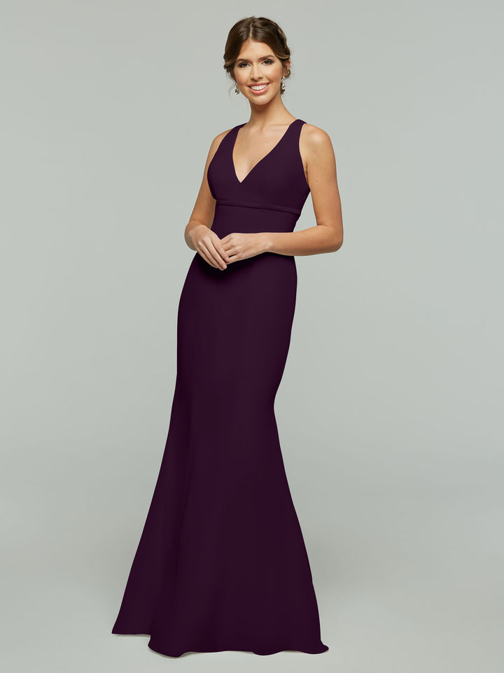 A woman smiles in the Avery Austin 80155, a sleeveless, floor-length deep purple chiffon dress with a V-neck and fit and flare silhouette, set against a plain light gray background.