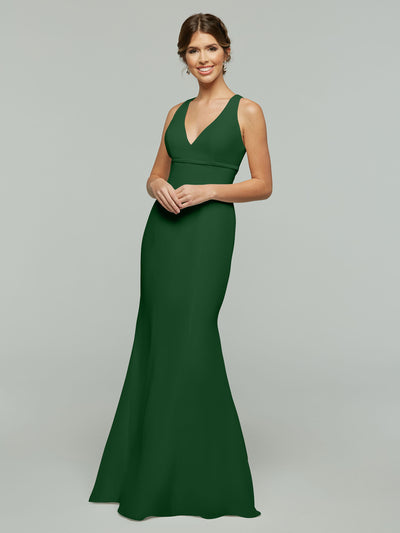 A woman with brown hair in a low updo smiles as she models Avery Austin’s 80155, a floor-length, sleeveless deep green chiffon dress with a V-neckline and fit and flare silhouette, posing against a plain light gray background.
