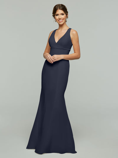 A woman in Avery Austin’s 80155, a sleeveless navy chiffon V-neck fit and flare gown, stands smiling against a plain light gray background.