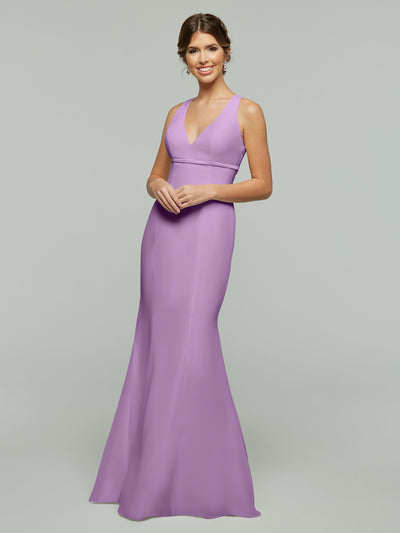 A woman with brown hair in a low updo smiles while wearing Avery Austin’s 80155: a long, sleeveless lilac chiffon dress with a fit and flare silhouette and V-neckline, posing against a plain light gray background.