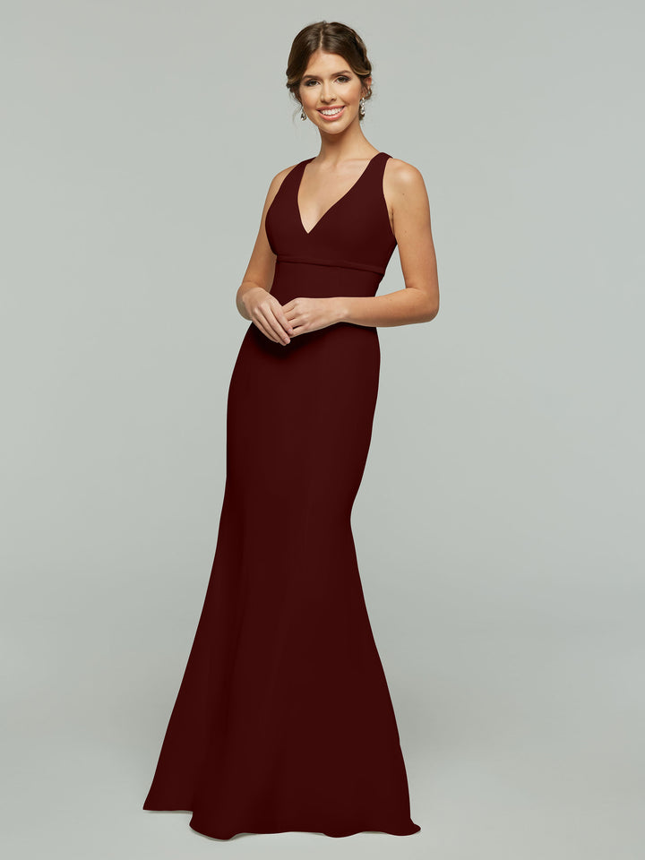 A woman wears Avery Austin’s 80155, a sleeveless, floor-length Merlot dress with a V-neckline and keyhole back, standing against a light gray background and smiling with hands gently clasped in front.