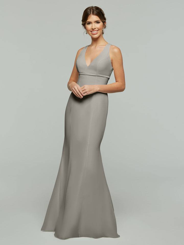 A woman with brown hair in an updo stands and smiles against a plain light gray background wearing the Avery Austin 80155 sleeveless fit and flare light gray V-neck gown.