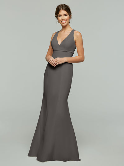 A woman wearing Avery Austin’s 80155 sleeveless, floor-length dark gray chiffon dress with a V-neckline and keyhole back stands against a light gray background, smiling with her hair in an updo.