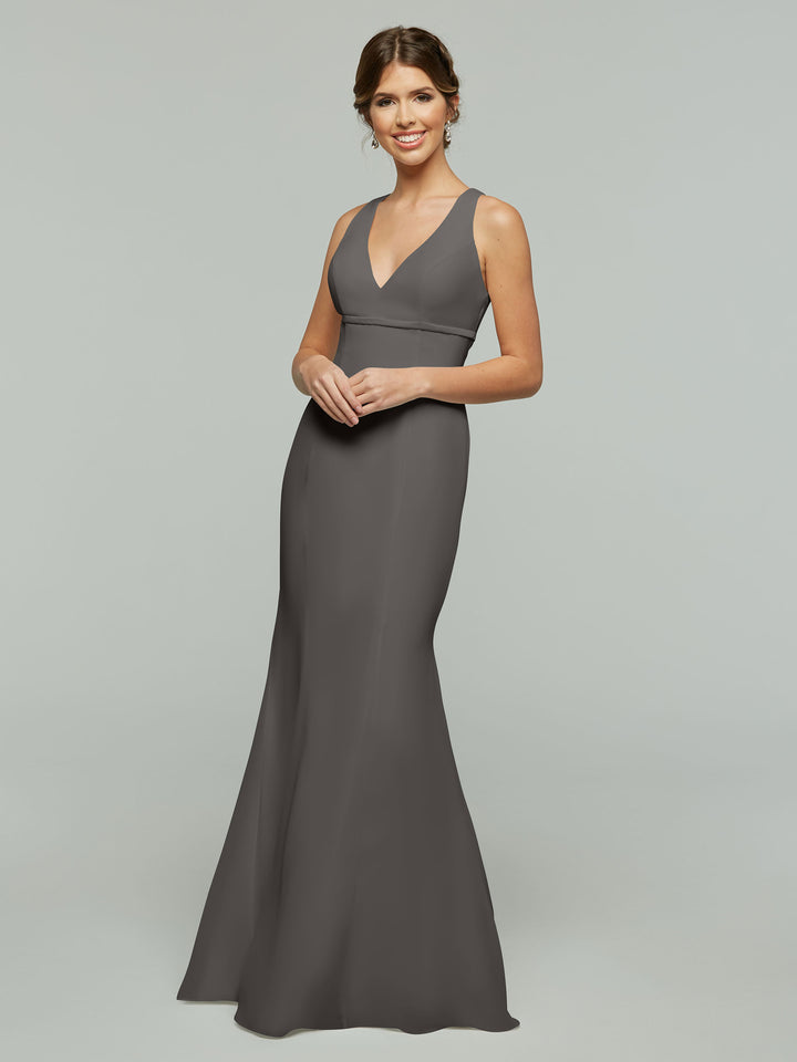 A woman smiles in an Avery Austin TH-80155 sleeveless, floor-length steel gray fit and flare gown with a V-neckline, standing against a plain light gray background.