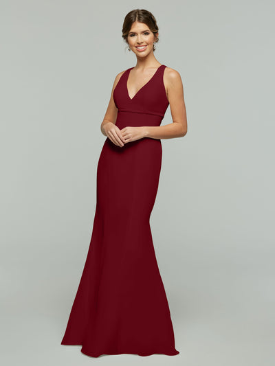 A woman smiles with her hands clasped in front of her, wearing Avery Austin style 80155—a sleeveless, V-neck, floor-length burgundy chiffon dress with a fit and flare silhouette—against a plain light gray background.